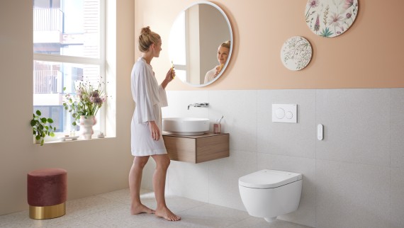 Egy fürdőszobában egy nő áll egy kedvező árú, mosdófunkcióval ellátott Geberit AquaClean Alba mellett Egy fürdőszobában egy nő áll egy kedvező árú, mosdófunkcióval ellátott Geberit AquaClean Alba mellett